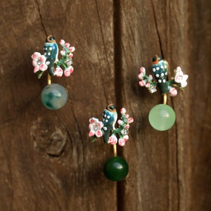 May include: Three pairs of earrings featuring a bird perched on a flowering branch. The birds are blue and white with orange accents. Each earring has a round, green stone at the base. The earrings are set against a wooden background.