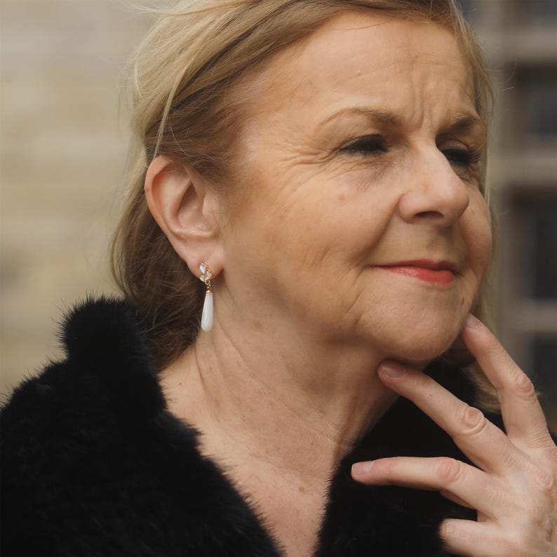 May include: A woman with blonde hair wearing a black fur coat and a pair of white teardrop earrings.