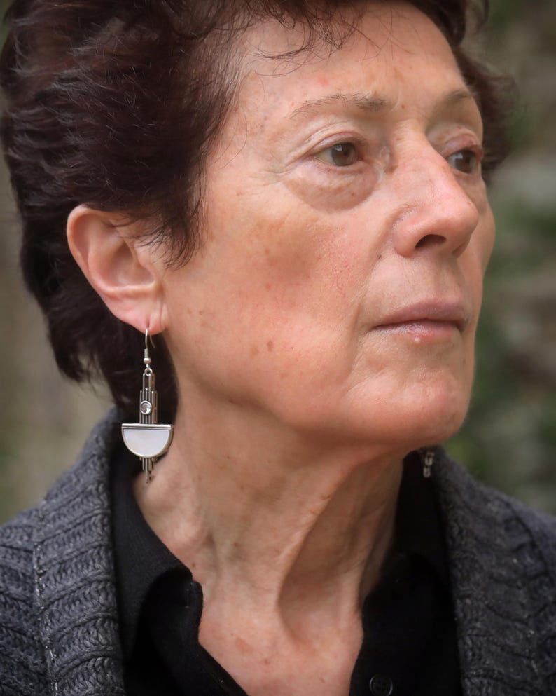May include: A silver-toned drop earring featuring a vertical rectangular bar with a small circular clear stone at the top and a semi-circular white inlay at the bottom, suspended from a simple silver-toned hook wire for pierced ears.