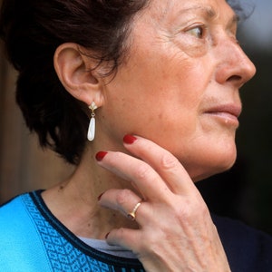 May include: A woman with short brown hair wearing a blue and black patterned shirt. She is wearing a gold earring with a white teardrop-shaped stone. She is also wearing a gold ring on her finger.