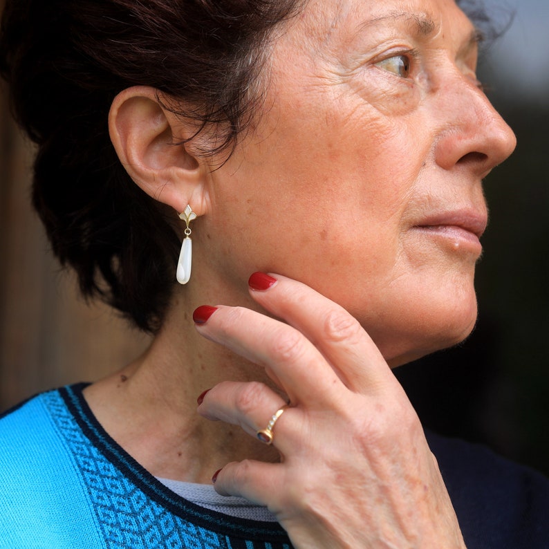 May include: A woman with short brown hair wearing a blue and black patterned shirt. She is wearing a gold earring with a white teardrop-shaped stone. She is also wearing a gold ring on her finger.