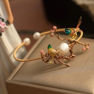 May include: A gold bracelet with a pearl and two birds perched on a branch with pink flowers. The birds are yellow, green, and blue.
