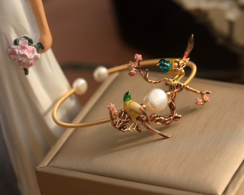 May include: A gold bracelet with a pearl and two birds perched on a branch with pink flowers. The birds are yellow, green, and blue.