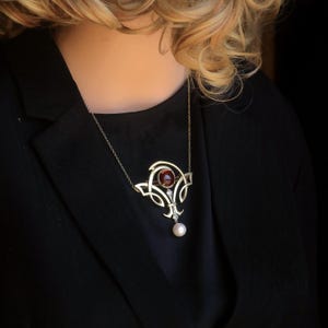 Peut inclure: Un collier doré avec un pendentif orné. Le pendentif présente une pierre précieuse rouge ronde au centre, un petit diamant et une perle pendante. Le collier est porté sur un vêtement noir. Le design est élégant.