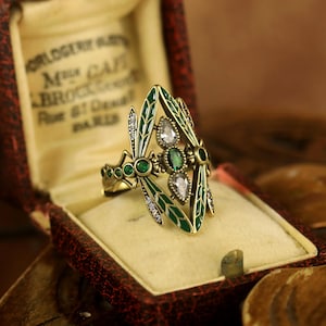 Könnte beinhalten: Ein goldfarbener Ring mit einem Libellen-Design. Die Flügel der Libelle sind aus grünem Emaille mit klaren und grünen Strasssteinen. Der Ring befindet sich in einer kleinen braunen Samtbox.