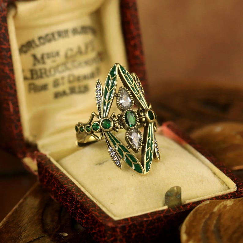 Pode incluir: Um anel dourado com um design de lib&eacute;lula. As asas da lib&eacute;lula s&atilde;o de esmalte verde com strass transparentes e verdes. O anel est&aacute; em uma pequena caixa de veludo marrom.