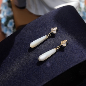 May include: A pair of white teardrop-shaped earrings with gold accents. The earrings have a delicate floral design and are made of mother-of-pearl.