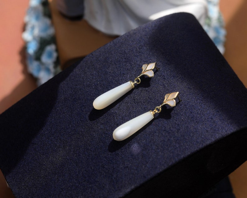May include: A pair of white teardrop-shaped earrings with gold accents. The earrings have a delicate floral design and are made of mother-of-pearl.