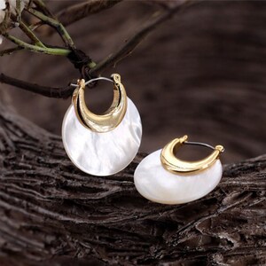 May include: A pair of gold hoop earrings with a white shell-like design. The earrings are on a wooden surface.