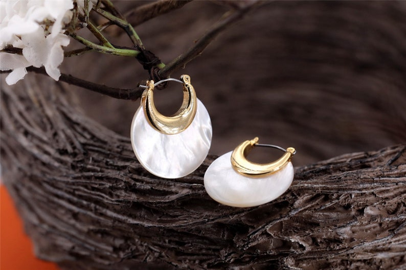 May include: A pair of gold hoop earrings with a white shell-like design. The earrings are on a wooden surface.