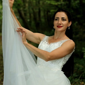 May include: A woman in a white wedding dress with lace detailing holds the veil up in front of her. She is standing in a forest setting.