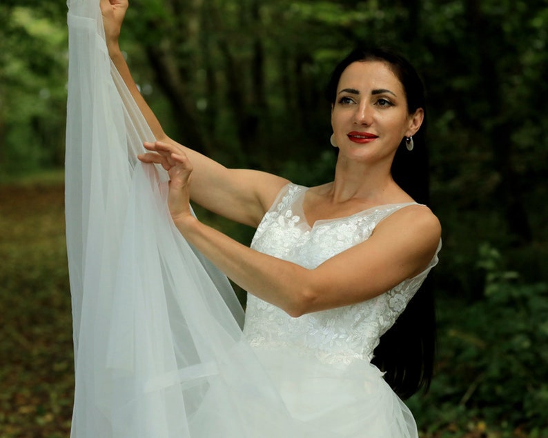 May include: A woman in a white wedding dress with lace detailing holds the veil up in front of her. She is standing in a forest setting.