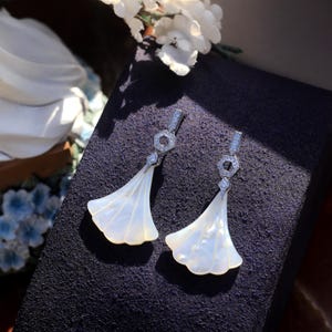 May include: A pair of elegant, fan-shaped earrings. Each earring features a silver-toned hexagonal top with small, clear gemstones and a flowing, white, shell-like pendant. The earrings are displayed on a dark purple surface, with white flowers in the background.