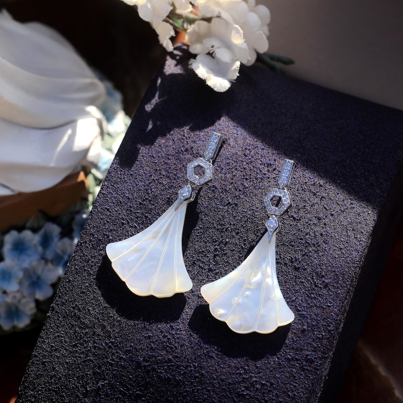 May include: A pair of elegant, fan-shaped earrings. Each earring features a silver-toned hexagonal top with small, clear gemstones and a flowing, white, shell-like pendant. The earrings are displayed on a dark purple surface, with white flowers in the background.