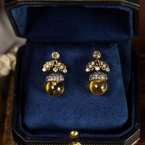 May include: A pair of gold acorn-shaped earrings with clear crystal accents. The earrings are in a blue velvet jewelry box.