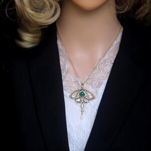 May include: A gold-toned pendant necklace with an emerald-green center stone, surrounded by clear crystals and a dangling pearl. The necklace is worn over a white lace top and a black blazer.