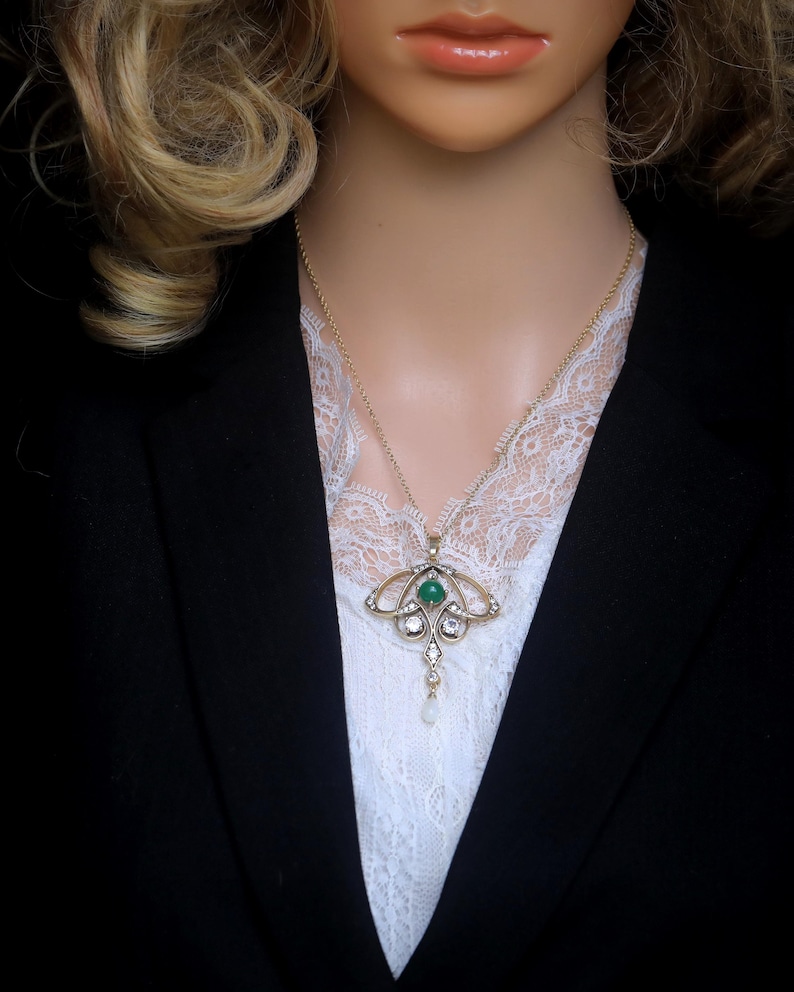 May include: A gold-toned pendant necklace with an emerald-green center stone, surrounded by clear crystals and a dangling pearl. The necklace is worn over a white lace top and a black blazer.