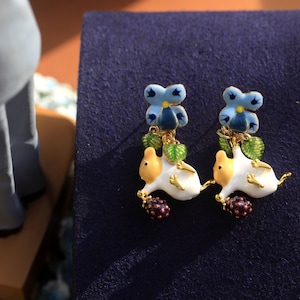 May include: A pair of earrings featuring a white ceramic mouse with a blue flower and green leaves. The earrings have a gold finish.