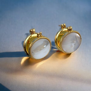 May include: A pair of gold hoop earrings with a white mother-of-pearl inlay. The earrings have a simple, elegant design.
