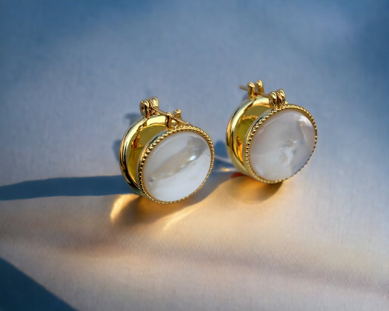 May include: A pair of gold hoop earrings with a white mother-of-pearl inlay. The earrings have a simple, elegant design.