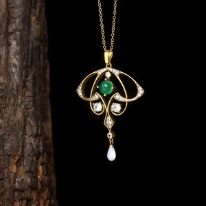 May include: A gold pendant necklace with an Art Nouveau design. The pendant features a central green gemstone, surrounded by clear gemstones and a dangling pear-shaped pearl. The necklace hangs on a delicate gold chain.