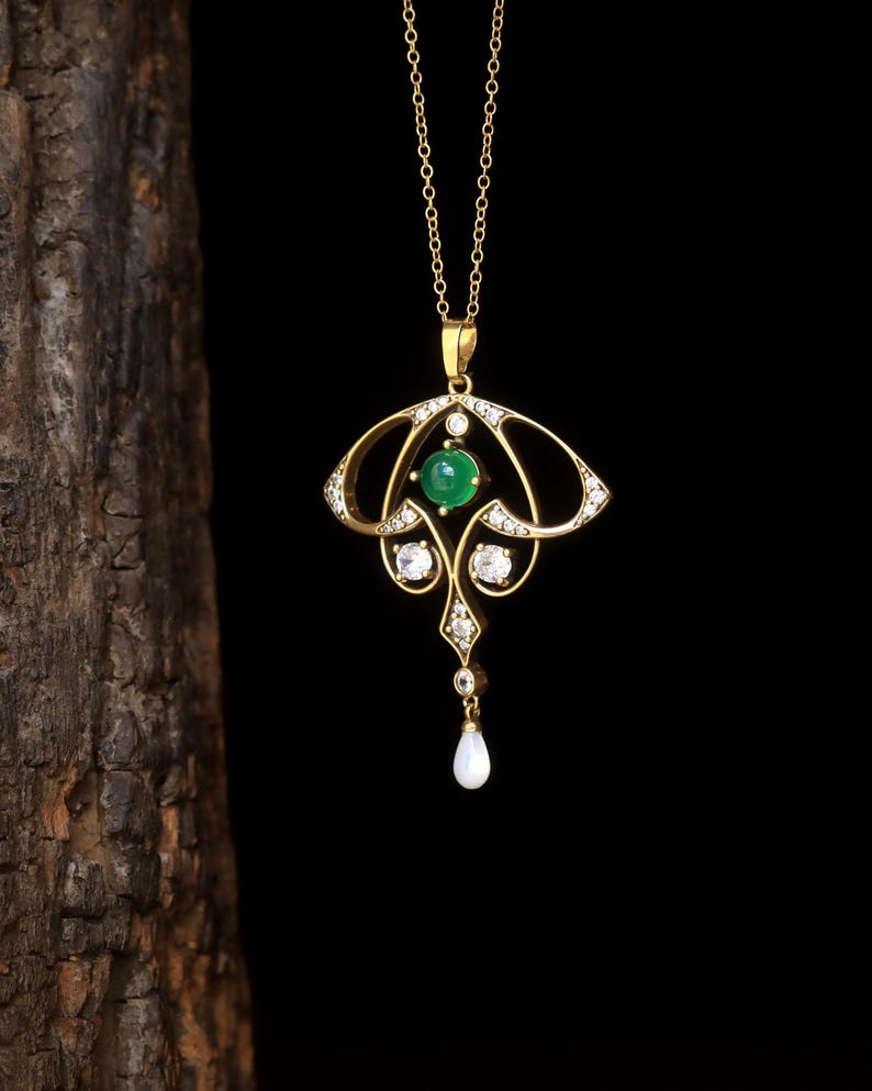 May include: A gold pendant necklace with an Art Nouveau design. The pendant features a central green gemstone, surrounded by clear gemstones and a dangling pear-shaped pearl. The necklace hangs on a delicate gold chain.
