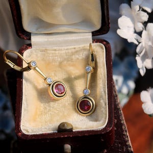 Op de afbeelding: Een paar gouden oorbellen met een beugel sluiting. Elke oorbel heeft een ronde, rode edelsteen omgeven door een gouden rand, en een kleine, ronde, heldere edelsteen. De oorbellen worden gepresenteerd in een bordeauxrode sieradendoos.