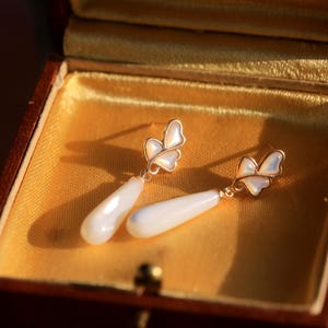 May include: A pair of gold butterfly earrings with white pearlescent teardrop dangles, displayed in a velvet-lined jewelry box.