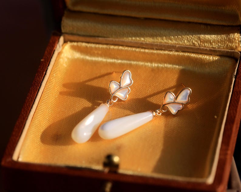 May include: A pair of gold butterfly earrings with white pearlescent teardrop dangles, displayed in a velvet-lined jewelry box.