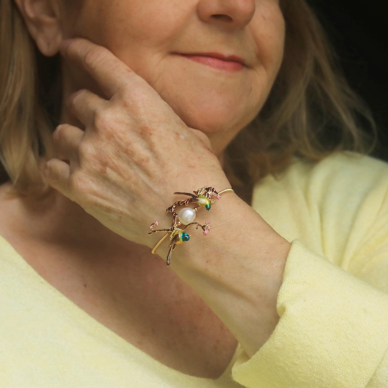 May include: A gold bracelet with a delicate design featuring two small birds perched on a branch with pink flowers and a pearl. The bracelet is on a person's wrist.