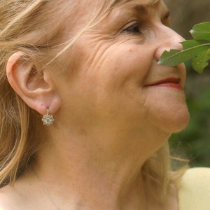 Puede incluir: Primer plano de la cara de una mujer, ella est&aacute; sonriendo y mirando hacia la derecha. Lleva un peque&ntilde;o pendiente de plata en forma de flor en la oreja izquierda. Su cabello es rubio y est&aacute; recogido hacia atr&aacute;s de su cara.