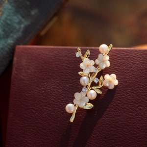 May include: A gold-tone brooch with a delicate floral design. The brooch features white flowers with pearl accents and clear rhinestones.