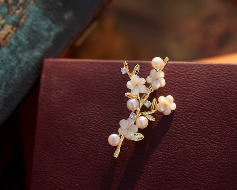 May include: A gold-tone brooch with a delicate floral design. The brooch features white flowers with pearl accents and clear rhinestones.