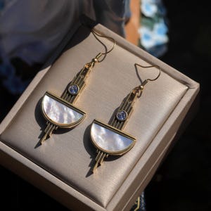 May include: A pair of gold-toned drop earrings featuring a semi-circular mother-of-pearl inlay at the base. Each earring has a vertical metal bar accented with a single clear round gemstone and a hook-style closure for secure attachment.
