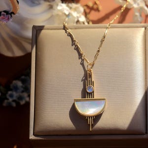 May include: A gold-toned necklace featuring a geometric pendant with a vertical bar, a small round clear stone, and a semi-circular inlay of iridescent white mother-of-pearl. The pendant hangs from a delicate gold-toned chain with textured links.
