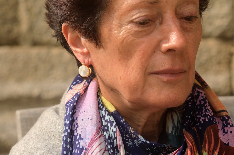 May include: A woman wearing a gold earring with a white pearl-like stone. The earring is a simple design with a small gold hoop.