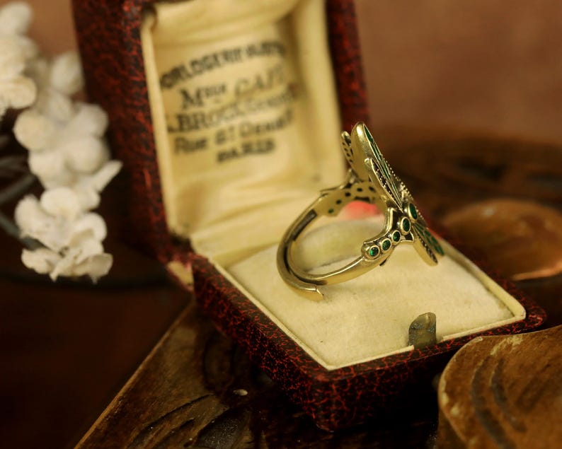 Pode incluir: Um anel de ouro com um design de lib&eacute;lula. As asas da lib&eacute;lula s&atilde;o decoradas com esmalte verde. O anel est&aacute; em uma caixa de veludo vermelho com a inscri&ccedil;&atilde;o "M. GATI, BROCKER, Rue et Ordeal, PARIS".