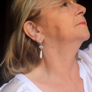 May include: A woman wearing a white shirt with silver earrings featuring a white pearl drop and a decorative fan-shaped design.