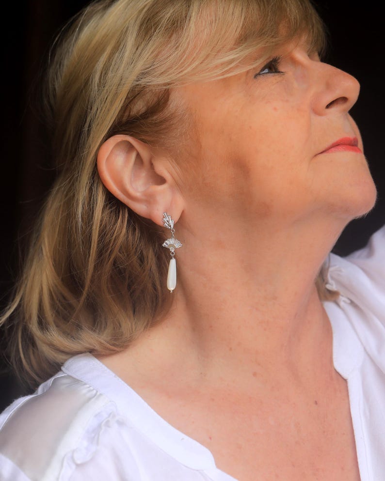 May include: A woman wearing a white shirt with silver earrings featuring a white pearl drop and a decorative fan-shaped design.