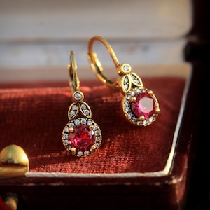 May include: A pair of gold-toned earrings with a central red gemstone surrounded by small, clear stones. The earrings have a lever-back closure and are displayed in a red velvet jewelry box. The design features a floral motif.