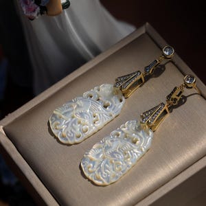 May include: A pair of white mother of pearl earrings with intricate floral designs. The earrings are gold-toned and feature clear crystal accents.