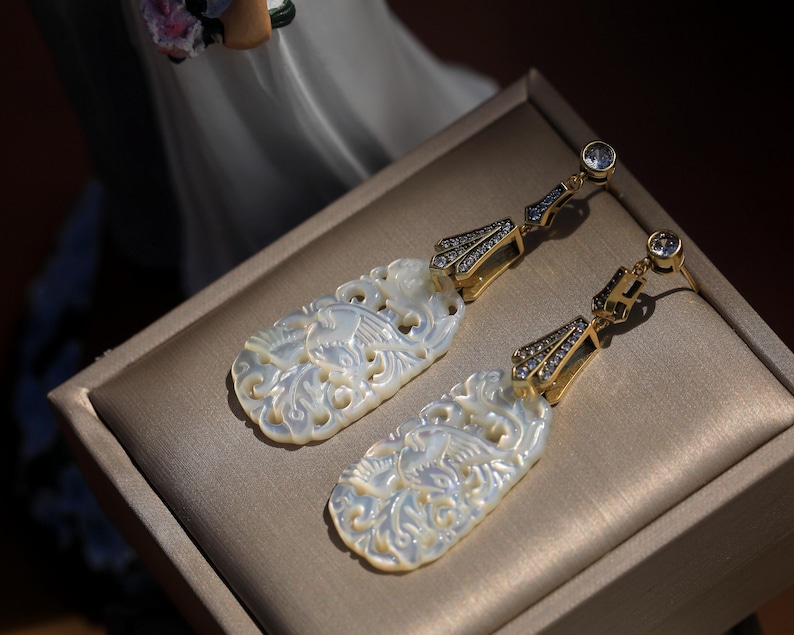 May include: A pair of white mother of pearl earrings with intricate floral designs. The earrings are gold-toned and feature clear crystal accents.