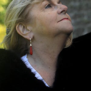 May include: A woman wearing a pair of gold earrings with a red teardrop-shaped coral pendant. The earrings are dangling. She is wearing a black fur coat and a white shirt. The woman has blonde hair and is looking up.