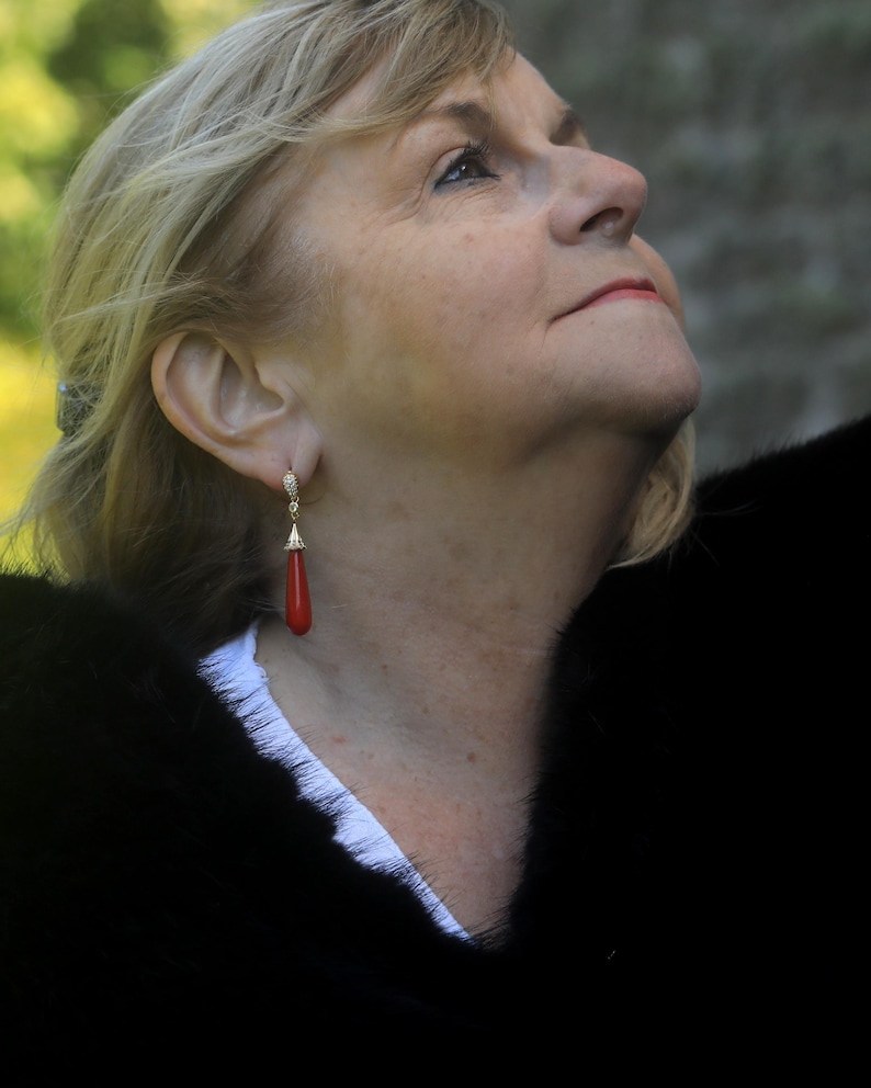 May include: A woman wearing a pair of gold earrings with a red teardrop-shaped coral pendant. The earrings are dangling. She is wearing a black fur coat and a white shirt. The woman has blonde hair and is looking up.