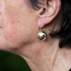 K&ouml;nnte beinhalten: Nahaufnahme eines Frauenohrs mit einem goldenen Creolenohrring mit einem runden, glatten, goldfarbenen Anh&auml;nger.