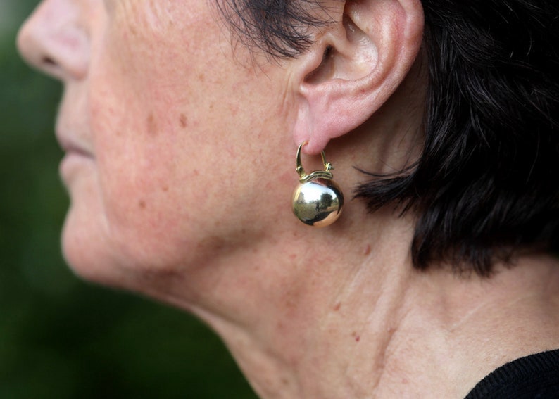 K&ouml;nnte beinhalten: Nahaufnahme eines Frauenohrs mit einem goldenen Creolenohrring mit einem runden, glatten, goldfarbenen Anh&auml;nger.