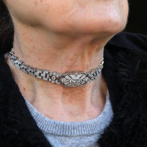 May include: A silver-toned choker necklace with a central rectangular design and intricate detailing. The necklace features numerous small, sparkling stones, creating a luxurious and elegant appearance. The design is symmetrical and classic.