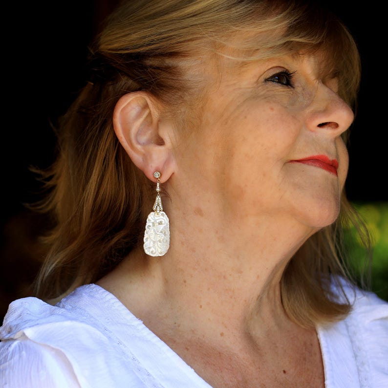 May include: A woman wearing white earrings with a textured, white, oval-shaped design. The earrings have a small, clear stone at the top.