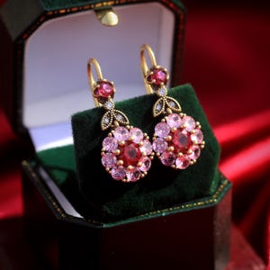 Könnte beinhalten: Ein Paar goldfarbene Ohrringe mit floralem Design, mit einem zentralen roten Edelstein, umgeben von rosa Edelsteinen. Die Ohrringe werden in einer grünen Samt-Schmuckschatulle vor rotem Hintergrund präsentiert.