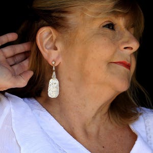 May include: A woman with blonde hair wearing a white shirt and silver dangle earrings with a floral design.